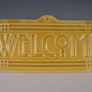 May include: A yellow ceramic welcome sign with a decorative border and raised lettering.