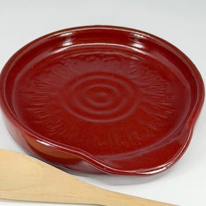 May include: A deep red ceramic spoon rest with a curved design. The spoon rest has a circular shape with a slight indentation for the spoon. A wooden spoon rests on the side of the spoon rest.