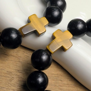 May include: Close-up of a beaded bracelet featuring large, black, round beads and cross-shaped accents. The crosses have a golden-brown, tiger-eye appearance. The bracelet is displayed on a white surface.