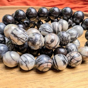 Puede incluir: Un brazalete de cuentas redondas de piedra natural, con cuentas negras, grises y crema, con vetas únicas. El brazalete se muestra sobre una superficie de madera.