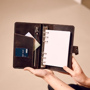 Op de afbeelding: Open bruin leren map met een ritsvak, kaartsleuf en een ringmechanisme voor het vasthouden van papier. Een blauwe Visa-kaart en een witte kaart zijn zichtbaar in de vakken. De map is open en toont blanco wit papier.