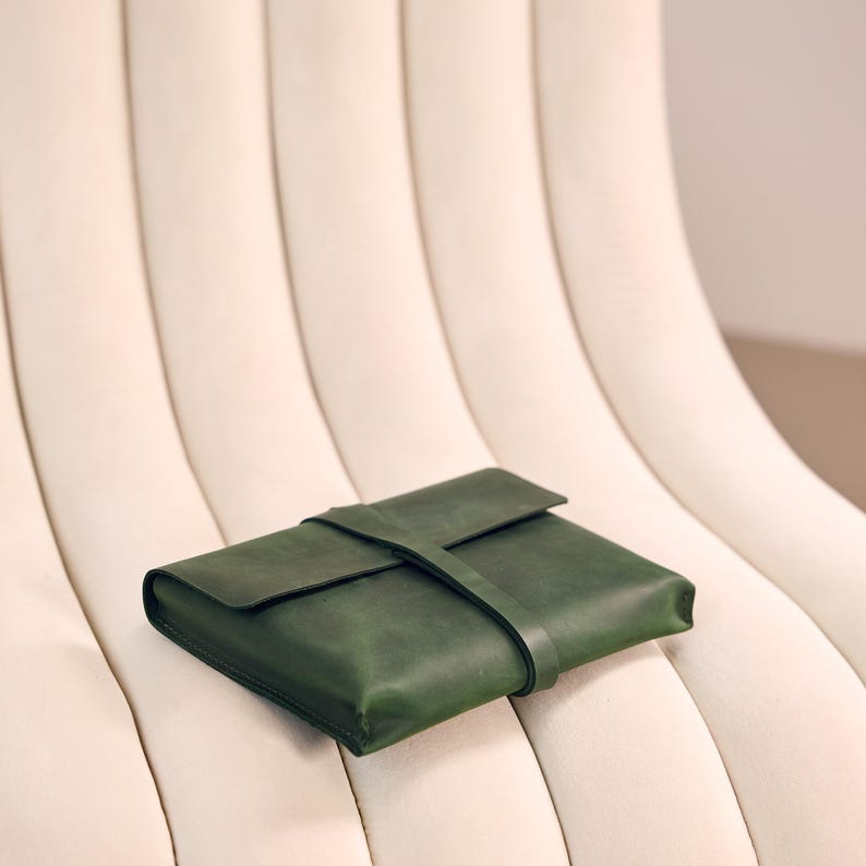 May include: A dark green leather folio with a flap and strap closure. The rectangular case rests on a cream-coloured, curved-back chair. The leather has a smooth texture and a matte finish.
