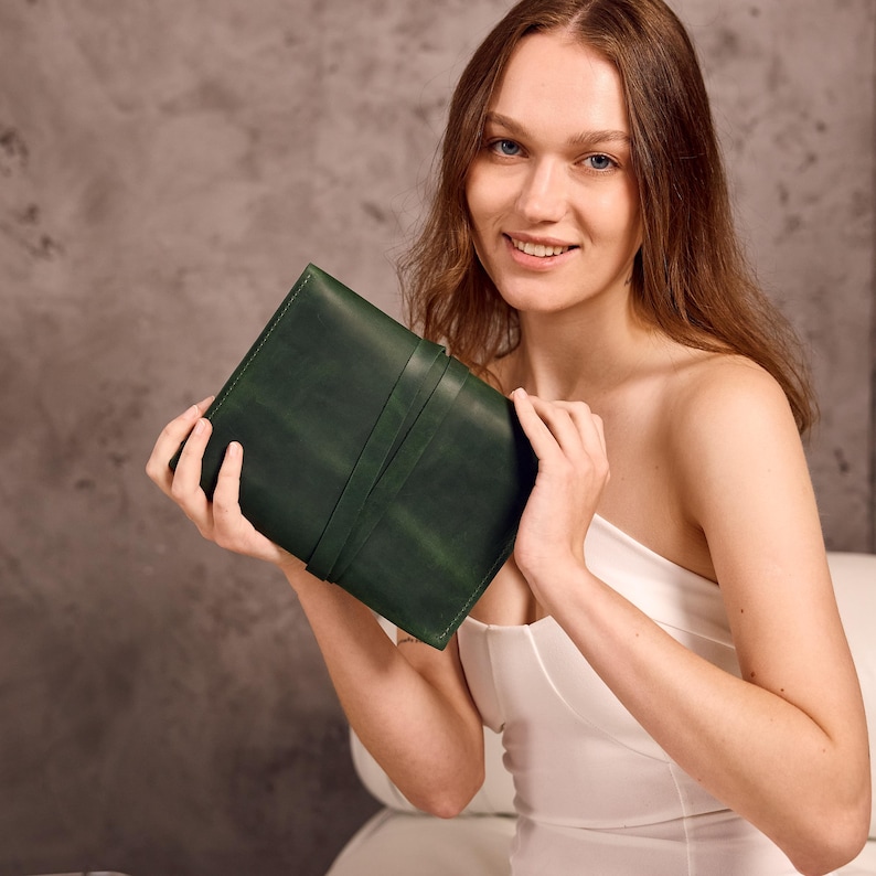 May include: A dark green leather journal with a strap closure. The journal is held open, showcasing its interior. The leather has a textured appearance. The journal is a rectangular shape.