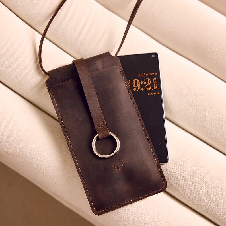 Może przedstawiać: Brązowe sk&oacute;rzane etui na telefon z paskiem, srebrnym pierścieniem i przeszyciami. Obok etui znajduje się czarny telefon z godziną 19:21.