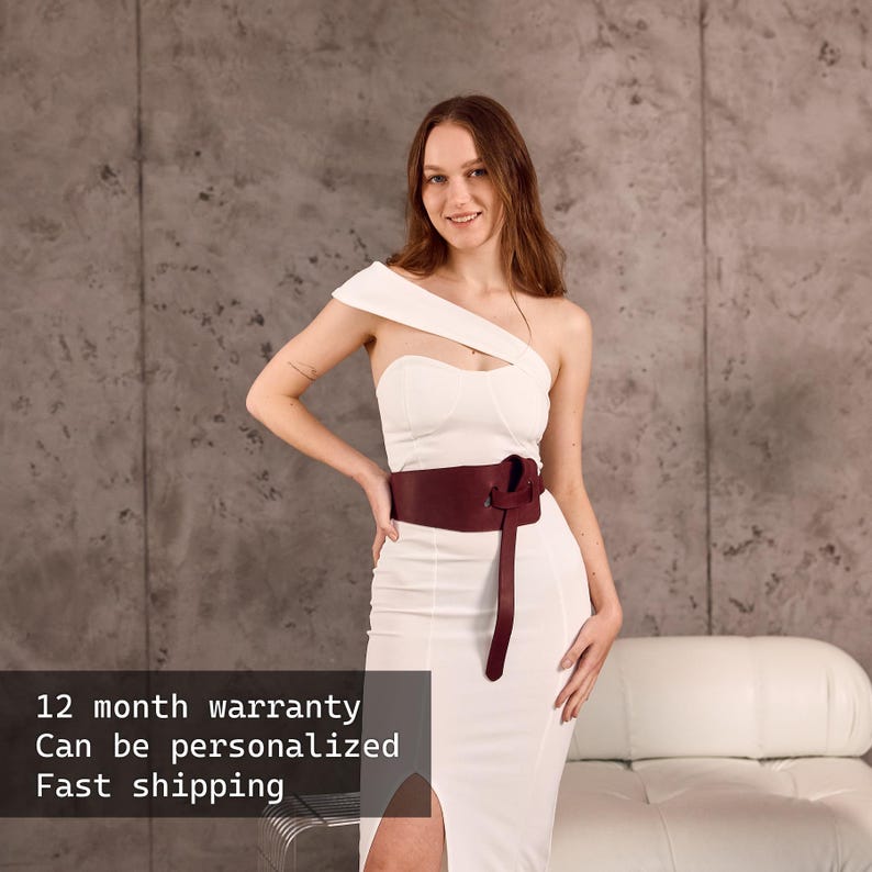 May include: A white, one-shoulder dress with a thigh-high slit, paired with a burgundy leather belt. The belt is tied in a knot at the front. The dress is worn by a person.