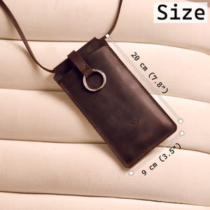 Może przedstawiać: Ciemnobrązowe sk&oacute;rzane etui na telefon z paskiem, o długości 20 cm (7,8") i szerokości 9 cm (3,5"). Posiada metalowy pierścień i mały element dekoracyjny. Widoczne jest słowo "Size".