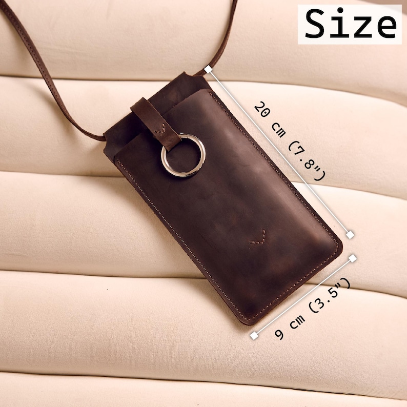 Może przedstawiać: Ciemnobrązowe sk&oacute;rzane etui na telefon z paskiem, o długości 20 cm (7,8") i szerokości 9 cm (3,5"). Posiada metalowy pierścień i mały element dekoracyjny. Widoczne jest słowo "Size".
