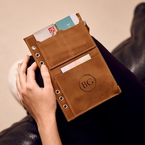 May include: A tan leather wallet with several card slots and a zipped section. The wallet is open, displaying cards and cash. The initials "BG" are embossed on the front. Metal eyelets are visible along the edge.