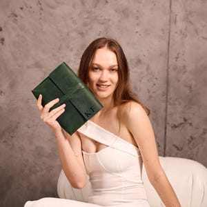 May include: A dark green leather journal with a wrap-around strap closure. The journal has the initials "KF" embossed on the front. The person is holding the journal in front of a grey background.