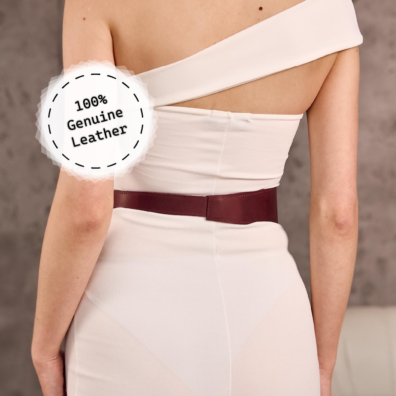 May include: A burgundy leather belt worn over a cream-colored dress. The belt is a solid color and appears to be made of genuine leather. The image includes the text "100% Genuine Leather".