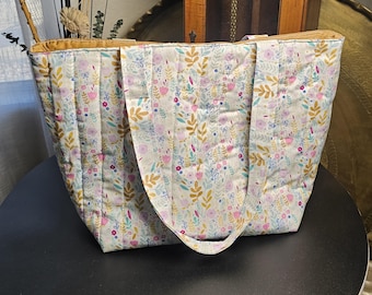 Puede incluir: Un bolso tote acolchado con un estampado floral. El bolso presenta un fondo crema con diseños florales y de hojas rosas, azules y dorados. El forro interior es dorado y el bolso tiene dos asas.