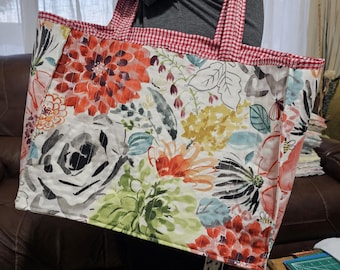 Könnte beinhalten: Eine große Tragetasche mit einem Blumenmuster in Rot-, Orange-, Gelb-, Grün- und Weißtönen. Der obere Rand und die Griffe sind rot-weiß kariert. Die Tasche ist aus gestepptem Stoff gefertigt.
