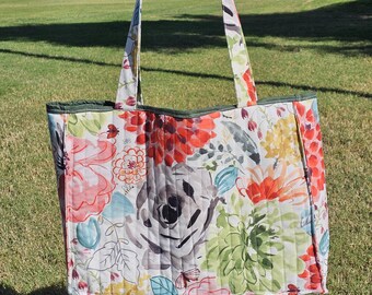 Könnte beinhalten: Eine gesteppte Tragetasche mit einem Blumenmuster in Rot-, Rosa-, Orange-, Grün- und Grautönen. Die Tasche hat eine grüne Einfassung und passende Blumenhenkel. Die Tasche wird vor einem grasbewachsenen Park hochgehalten.
