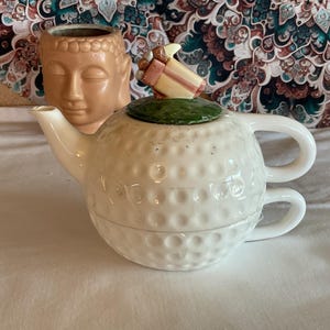 Könnte beinhalten: Weißes Keramik-Teeservice mit Golfball-Design. Die Teekanne hat einen grünen Deckel und ein dekoratives Geschenk oben. Im Hintergrund befindet sich ein kleiner, hellbrauner Buddha-Kopf.