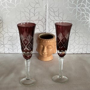 Könnte beinhalten: Zwei rubinrote Kristall-Champagnerflöten mit aufwendigen Ätzmustern. Die Flöten haben klare Stiele und Böden. Eine kleine, hellbraune Buddha-Kopf-Statue befindet sich zwischen den Flöten. Der Hintergrund ist ein silbernes und weißes geometrisches Muster.