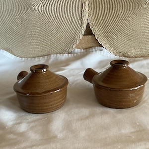 May include: Two small, brown ceramic pots with lids and handles. The pots have a rounded shape and a dark brown glaze. They are on a white surface, with two circular woven placemats in the background.