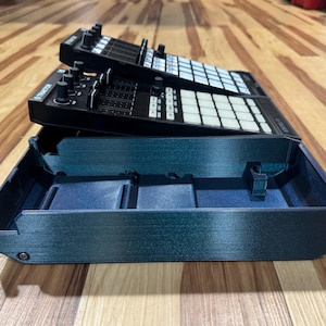 May include: Black DJ controller with white buttons and knobs, stacked on a dark teal tray. The controller has the word "TRAKTOR" printed on it. The tray has a rectangular shape with a low profile.