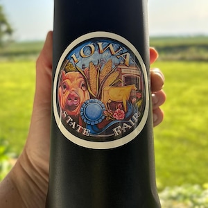 May include: A matte black cylindrical water bottle featuring a circular sticker with the text IOWA STATE FAIR. The sticker displays a pig, ears of corn, a cow, a blue ribbon, and pink flowers in a colorful, detailed illustration style.