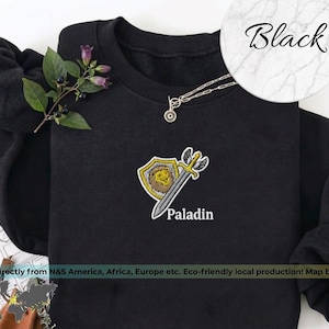 Può includere: Felpa nera con un design ricamato con uno scudo con un leone, una spada alata e la parola "Paladin". Sono visibili anche una collana d'argento e un rametto di verde. La parola "Black" è scritta in un cerchio.