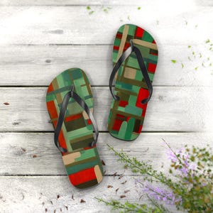 Op de afbeelding: Een paar teenslippers met een kleurrijk abstract ontwerp. De sandalen hebben een patroon van groene, rode en bruine rechthoeken en vierkanten. De bandjes zijn zwart. De teenslippers worden gepresenteerd op een wit houten oppervlak.