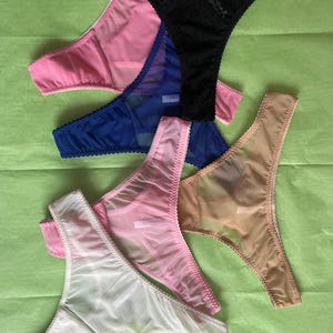 May include: A collection of sheer thong underwear in various colors, including black, pink, blue, white, and tan. The lingerie features delicate stitching and a minimalist design. The garments are arranged on a green surface.