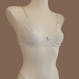May include: A white ribbed lingerie set consisting of a triangle bra and matching thong. Both pieces feature thin adjustable straps and a small, delicate pink rosebud accent centred at the neckline of the bra and the front of the thong.
