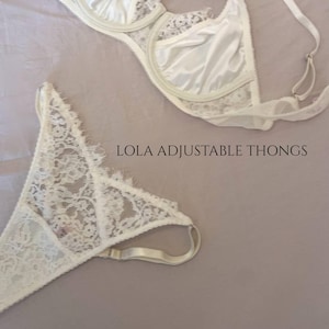 May include: A white lace lingerie set comprising a bra and adjustable thong underwear. The bra features underwire cups with lace detailing, and the thong is crafted from matching white lace. The text LOLA ADJUSTABLE THONGS is clearly visible.