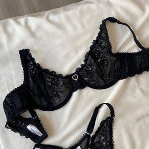 Könnte beinhalten: Schwarzes Spitzen-BH und passendes String-Set. Die Dessous weisen zarte florale Spitzenmuster und kleine herzförmige Verzierungen auf. Das Set wird auf einem weißen Stoffhintergrund präsentiert.