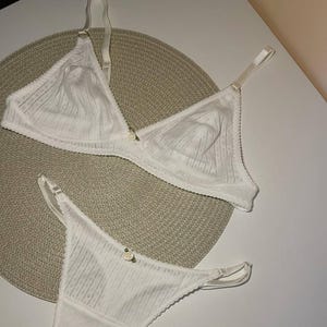 May include: A white lingerie set comprising a triangle bra and thong. The bra has adjustable straps and a small floral detail. Both items are made from a textured, ribbed fabric with delicate lace edging. The set is presented on a neutral-toned mat.