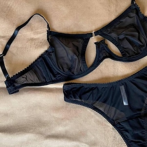 May include: Black lingerie set featuring a sheer bra with a unique cut-out design and adjustable straps. The set includes matching low-rise underwear. The fabric appears to be a soft, delicate material.