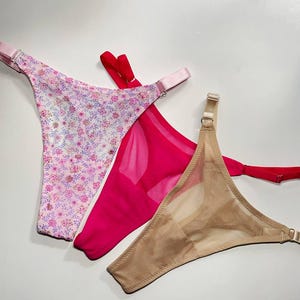 Può includere: Tre perizomi con spalline regolabili e cursori in metallo. Un capo è rosa chiaro con un motivo floreale, uno è fucsia a tinta unita con inserti in rete trasparente e uno è beige a tinta unita con inserti in rete trasparente.