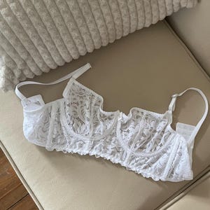 May include: A white lace underwire bra featuring a floral pattern, adjustable shoulder straps with small decorative embellishments, and a hook-and-eye back closure. The garment is constructed from sheer white lace fabric with scalloped edges.