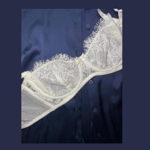 May include: A white lace underwire bra with scalloped edges and a small center bow, layered over a dark blue button-down shirt. The bra features intricate floral patterns in the lace and adjustable straps, contrasting against the smooth fabric.