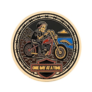 Pode incluir: Um broche metálico circular com um esqueleto a conduzir uma mota ao longo de uma praia. O design inclui uma borda de arame farpado, uma onda azul e um escudo vermelho na parte inferior com o texto dourado ONE DAY AT A TIME em moldura preta.