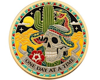 Op de afbeelding: Een ronde goudkleurige metalen munt met een kleurrijk emaille ontwerp van een schedel, een groene cactus, een kronkelende slang en een rode roos tegen een woestijnlandschap. De tekst ONE DAY AT A TIME staat op een banner onderaan de munt.