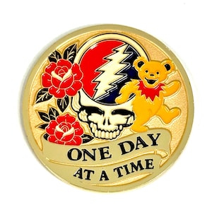May include: A round, gold-colored coin featuring a skull with a lightning bolt, a dancing bear, and red roses. The text "ONE DAY AT A TIME" is displayed on a banner. The coin has a gold border.