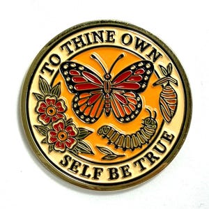Puede incluir: Una moneda redonda de metal dorado con un diseño de mariposa, oruga y crisálida. La moneda presenta el texto "TO THINE OWN SELF BE TRUE" en un patrón circular alrededor del borde. La mariposa es naranja, roja y negra.