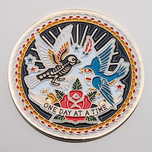 May include: A round, gold-colored metal coin with a detailed design. The coin features two birds, one black with a skeleton design and the other blue, facing each other. A red rose and the words "ONE DAY AT A TIME" are also included.