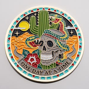 Könnte beinhalten: Ein rundes, farbenfrohes Metallabzeichen mit Wüstenmotiv. Es zeigt einen Schädel, einen Kaktus und Schlangen, mit den Worten "ONE DAY AT A TIME" auf einem Banner. Das Design umfasst rote, gelbe, grüne und blaue Akzente.