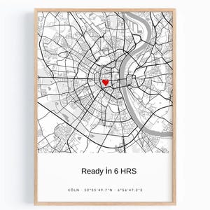 Peut inclure: Une carte en noir et blanc encadrée de Cologne, en Allemagne, avec un cœur rouge indiquant un emplacement. Le texte "Ready in 6 HRS" est en dessous de la carte, avec les coordonnées de la ville. Le cadre est en bois clair.