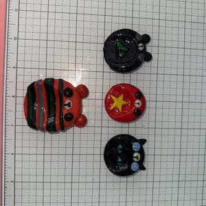 Puede incluir: Una colecci&oacute;n de pines de arcilla hechos a mano con dise&ntilde;os de animales. Los pines incluyen un oso a rayas, un oso con un tri&aacute;ngulo verde, un c&iacute;rculo rojo con una estrella amarilla y un gato negro con ojos azules.