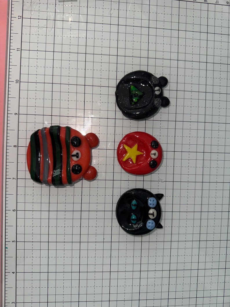 Puede incluir: Una colecci&oacute;n de pines de arcilla hechos a mano con dise&ntilde;os de animales. Los pines incluyen un oso a rayas, un oso con un tri&aacute;ngulo verde, un c&iacute;rculo rojo con una estrella amarilla y un gato negro con ojos azules.