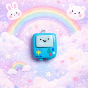 Puede incluir: Un dije azul claro con forma de consola de videojuegos retro, con una pantalla blanca y una cara sonriente. El dije tiene un lazo plateado en la parte superior para colgarlo. El fondo presenta un arcoíris, nubes y personajes de dibujos animados.