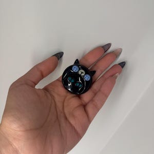 Puede incluir: Broche negro con forma de gato con ojos de bot&oacute;n azules y blancos y ojos turquesa. El broche es redondo y brillante, con un dise&ntilde;o de cara de gato. El broche se sostiene en una mano sobre un fondo blanco.