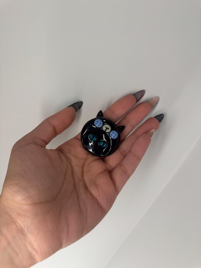 Puede incluir: Broche negro con forma de gato con ojos de bot&oacute;n azules y blancos y ojos turquesa. El broche es redondo y brillante, con un dise&ntilde;o de cara de gato. El broche se sostiene en una mano sobre un fondo blanco.