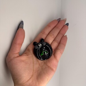 Puede incluir: Un colgante redondo negro con un contorno blanco y un dise&ntilde;o de tri&aacute;ngulo verde en el centro. El colgante tiene peque&ntilde;os adornos grises y se sostiene en una mano.