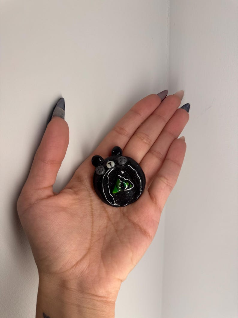 Puede incluir: Un colgante redondo negro con un contorno blanco y un dise&ntilde;o de tri&aacute;ngulo verde en el centro. El colgante tiene peque&ntilde;os adornos grises y se sostiene en una mano.