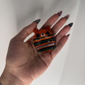 Puede incluir: Una peque&ntilde;a figura con forma de oso naranja con ojos de bot&oacute;n negros y una bufanda a rayas verdes, naranjas y grises. El oso tiene orejas peque&ntilde;as y redondeadas y una nariz negra. La figura probablemente est&eacute; hecha de vidrio o un material similar.