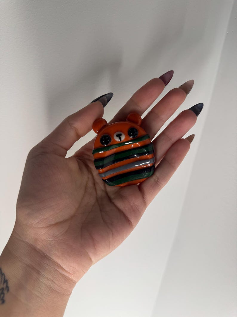 Puede incluir: Una peque&ntilde;a figura con forma de oso naranja con ojos de bot&oacute;n negros y una bufanda a rayas verdes, naranjas y grises. El oso tiene orejas peque&ntilde;as y redondeadas y una nariz negra. La figura probablemente est&eacute; hecha de vidrio o un material similar.