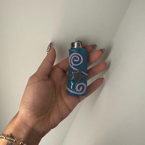 Può includere: Un accendino turchese con la parte superiore argentata. È decorato con un motivo a spirale rosa e una stella grigia. L'accendino è tenuto in una mano.